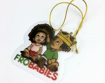 May include: A clear plastic ornament with a gold string for hanging. The ornament features a cartoon illustration of two Black children with Afro hairstyles. The girl is wearing a pink shirt and red shorts, and the boy is wearing a green shirt and blue shorts. The boy is wearing a gold crown. The text "FRO BABIES" is printed in red and green on the ornament.