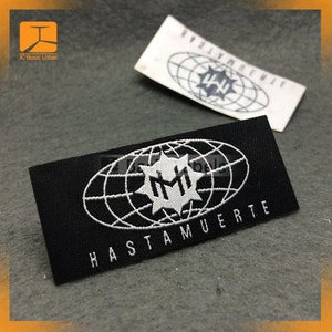 May include: Black woven label with white text and a logo. The logo is a stylized globe with a star in the center. The text reads "HASTA MUERTE" and "HM".
