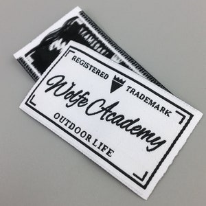 May include: White woven label with black text and a black triangle with a crown. The text reads "Registered Trademark", "Wolfe Academy", and "Outdoor Life".