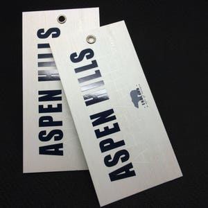 Pode incluir: Duas etiquetas retangulares brancas com textura de madeira e texto preto que diz "Aspen Hills" com um pequeno logotipo de urso. O texto &eacute; ligeiramente elevado e tem um acabamento brilhante.
