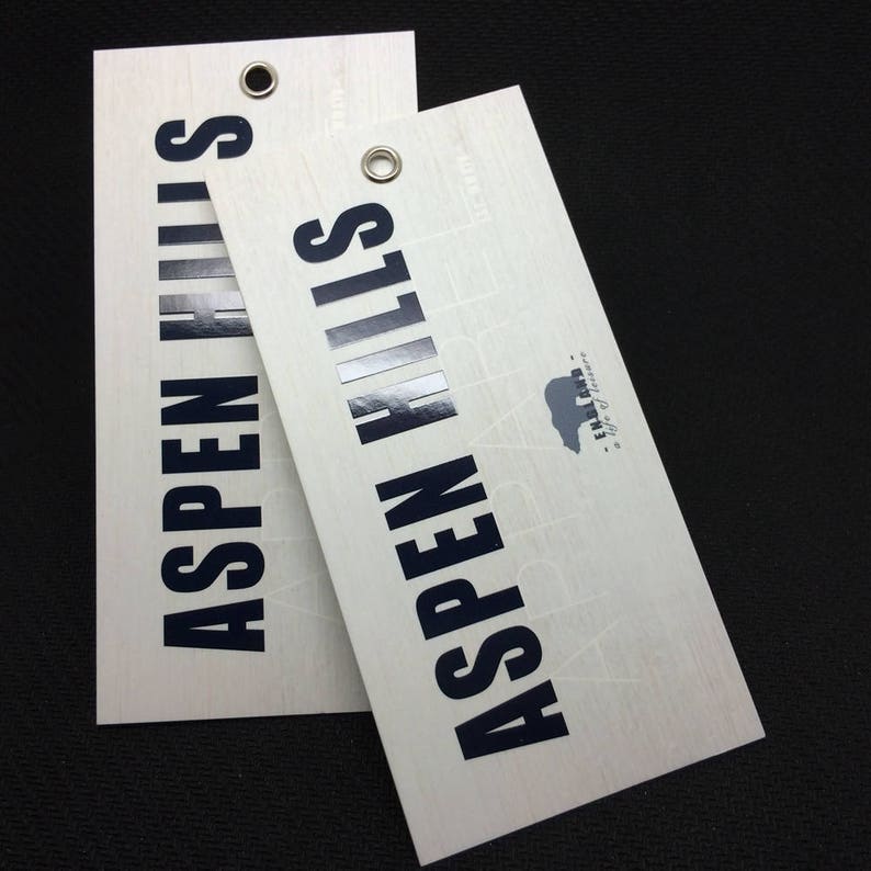 Pode incluir: Duas etiquetas retangulares brancas com textura de madeira e texto preto que diz "Aspen Hills" com um pequeno logotipo de urso. O texto &eacute; ligeiramente elevado e tem um acabamento brilhante.