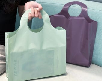 Op de afbeelding: Twee draagtassen, waarvan één door een persoon wordt vastgehouden. De eerste tas is lichtgroen en de tweede is dieppaars. Beide tassen hebben een ronde handgreep en het woord "BEYOND" is zichtbaar op de groene tas.