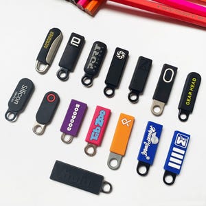 Könnte beinhalten: Eine Sammlung von Reißverschlussanhängern in verschiedenen Farben und Designs. Einige Anhänger sind schwarz mit weißem Text, andere sind lila, orange und blau mit verschiedenen Logos und Texten. Die Anhänger haben Metallschlaufen zur Befestigung.