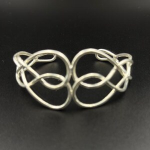 Peut inclure: Un bracelet manchette de couleur argentée présentant un design ajouré composé de formes de fils entrelacées et bouclées qui forment un motif continu et symétrique sur le devant de la pièce, créant une apparence légère et aérée.