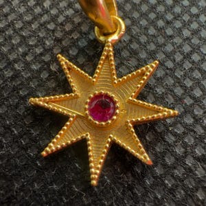 Könnte beinhalten: Goldfarbener Sternanhänger mit einem zentralen, runden, roten Edelstein. Der Anhänger hat acht Zacken, jede mit einem Perlenrand. Die Öse ist eine einfache Schlaufe.