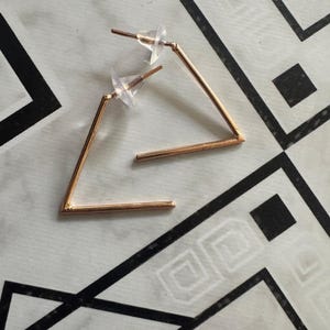 Pode incluir: Um par de brincos triangulares geométricos dourados. Os brincos têm um design simples, com um pino e fecho. Os brincos são exibidos em uma superfície branca com um padrão geométrico em preto e branco.