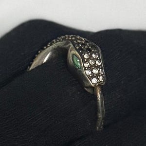 May include: A silver snake ring with a detailed head. The snake's head is encrusted with small, clear crystals and black stones. The snake has a green eye and a curved tongue. The ring is set against a black background.