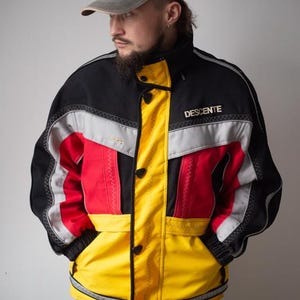 May include: A black, red, and yellow ski jacket with the word "DESCENTE" printed on the chest. The jacket has a high collar, button closures, and reflective silver stripes. A gray baseball cap is worn.
