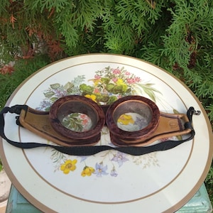Puede incluir: Gafas steampunk vintage con lentes circulares marrón oscuro y montura de cuero marrón claro. Las gafas tienen una correa negra ajustable. Se encuentran sobre un fondo con estampado floral.
