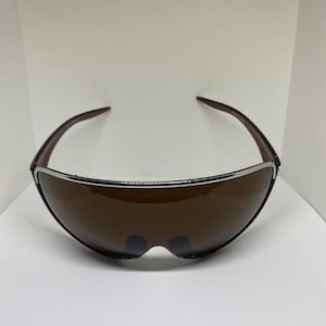 Op de afbeelding: Een zonnebril in shield-stijl met een enkele doorlopende bruin getinte lens en een dun zilverkleurig metalen frame dat de bovenrand omlijnt. De donkerbruine pootjes buigen naar binnen voor een strak en gestroomlijnd uiterlijk.