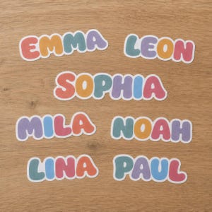 May include: A collection of six die-cut vinyl stickers featuring the names EMMA, LEON, SOPHIA, MILA, NOAH, LINA, and PAUL. Each letter is rendered in a rounded, bubble-style font with a multi-colored palette including red, yellow, teal, and purple.
