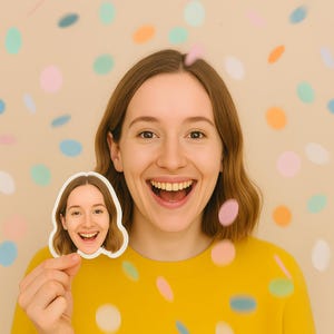Op de afbeelding: Een vrouw in een gele trui lacht breeduit en houdt een sticker van haar eigen gezicht vast. De sticker heeft een witte rand. De achtergrond heeft een beige muur met kleurrijke confetti-achtige vormen.