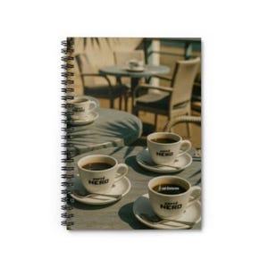 May include: Spiral-bound notebook featuring a photograph of several cups of coffee on saucers, with the text "caffè NERO" on the cups. The background shows outdoor seating at a cafe, with tables and chairs.