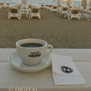 May include: A white ceramic cup of coffee with the text "CAFFE NERO" sits on a saucer next to a folded napkin with a logo. The scene is set on a white wooden table with a beach and umbrellas in the background.