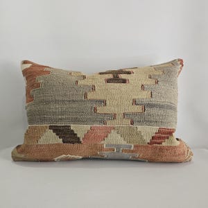 Könnte beinhalten: Rechteckiges Zierkissen mit einem geometrischen Muster in Beige, Grau, Braun und Rosa. Das Kissen hat eine gewebte Textur und eine neutrale Farbpalette, die für verschiedene Einrichtungsstile geeignet ist.