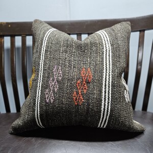 Pode incluir: Almofada decorativa quadrada com textura tecida cinza escuro. Apresenta listras brancas verticais e padrões florais bordados em laranja, roxo e amarelo. A almofada está colocada em uma cadeira marrom escura. Mede aproximadamente 40 cm x 40 cm.