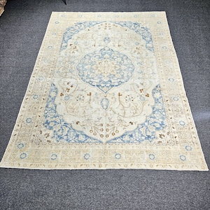 May include: Rectangular area rug with an intricate floral design. The rug features a cream-colored background with a large central medallion and border in shades of blue and brown. The rug is made of a soft material.