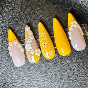 May include: Five long, almond-shaped artificial nails. The nails are painted in shades of yellow and pink, with floral and dot designs. Some nails feature small, white flower embellishments. The nails are arranged on a clear plastic holder against a black background.