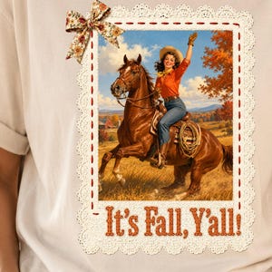 May include: A cream-colored t-shirt featuring a rectangular graphic of a person riding a brown horse in a field. The graphic is framed by a white lace border, a small floral bow, and the orange text "It's Fall, Y'all!" printed at the bottom.