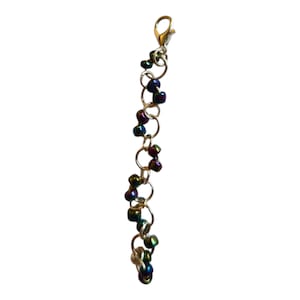 Peut inclure: Un charm argenté avec des perles irisées et des maillons circulaires. Le charm est doté d'un fermoir mousqueton en haut et est composé de petites perles colorées dans les tons de violet, vert et bleu. Les perles sont reliées par des anneaux argentés.