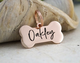 May include: A rose gold dog tag in the shape of a bone. The tag has the name "Oakley" in black script. The tag is attached to a rose gold ring.