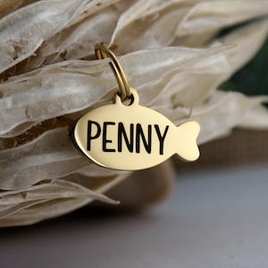 May include: A gold-colored pet tag in the shape of a fish, with the name "PENNY" in black capital letters. The tag is attached to a gold-colored ring. The background is blurred, with beige and green tones.
