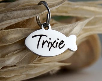 May include: A white, fish-shaped pet tag with the name "Trixie" in black script. The tag is attached to a silver-colored ring. The background features dried, beige-colored plant matter.