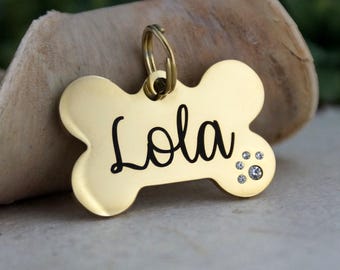 May include: Gold bone-shaped dog tag with the name "Lola" engraved in black lettering and a paw print made of clear rhinestones.