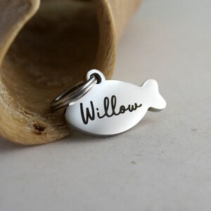 May include: A silver-colored pet tag in the shape of a fish. The name "Willow" is written in black cursive script across the tag. A small metal ring is attached to the top for easy attachment to a pet's collar.