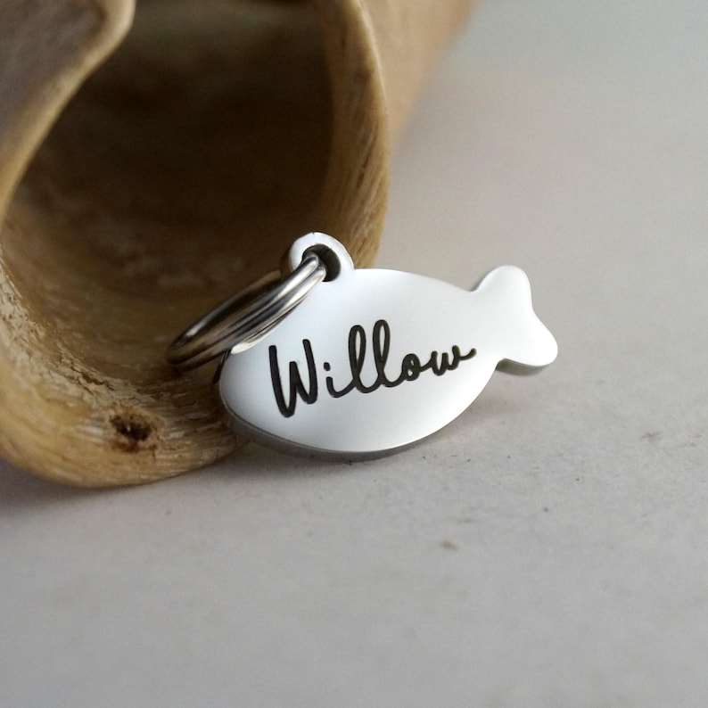 May include: A silver-colored pet tag in the shape of a fish. The name "Willow" is written in black cursive script across the tag. A small metal ring is attached to the top for easy attachment to a pet's collar.
