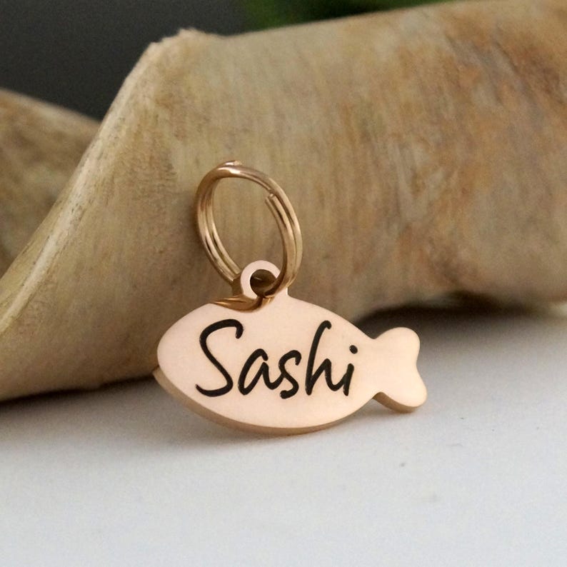 May include: A rose gold pet tag in the shape of a fish. The tag has a ring for attachment and the name "Sashi" in black script. The tag is on a light brown surface.