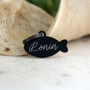 May include: A black, fish-shaped pet tag with the name "Ronin" in a cursive font. The tag has a small ring at the top for attachment to a collar. The background is a blurred, light-colored wooden surface.