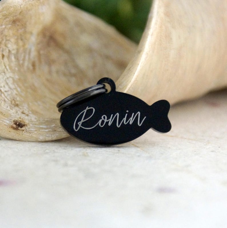 May include: A black, fish-shaped pet tag with the name "Ronin" in a cursive font. The tag has a small ring at the top for attachment to a collar. The background is a blurred, light-colored wooden surface.