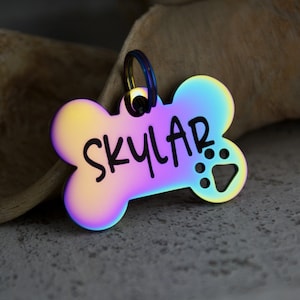 May include: A rainbow iridescent dog tag in the shape of a bone with the name "SKYLAR" engraved in black lettering and a paw print cutout.