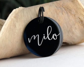 May include: A black, circular pet tag with the name "milo" in white cursive script. The tag has a black metal ring for attachment. The background is a piece of light-colored wood.