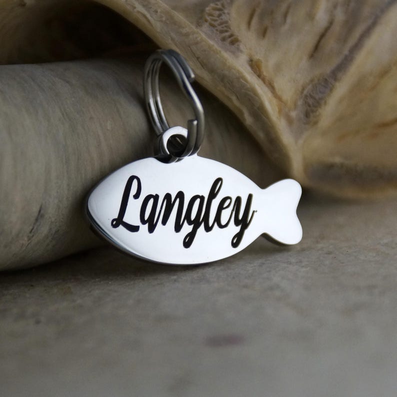 May include: A shiny, silver-colored pet tag in the shape of a fish. The name "Langley" is written in a cursive font across the tag. A small metal ring is attached at the top for easy attachment to a collar.
