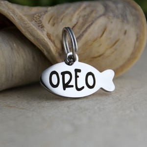 May include: A silver-colored pet tag in the shape of a fish. The tag has the name "OREO" engraved in bold, black letters. The tag is attached to a silver ring.