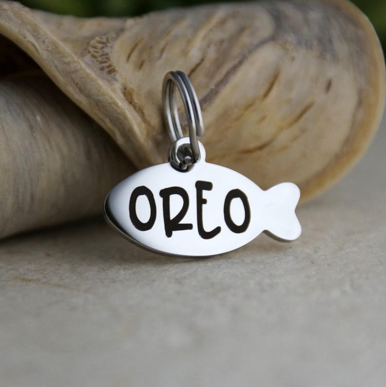 May include: A silver-colored pet tag in the shape of a fish. The tag has the name "OREO" engraved in bold, black letters. The tag is attached to a silver ring.