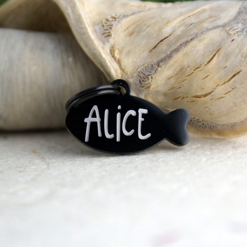May include: A black, fish-shaped pet tag with the name "Alice" in white, sans-serif font. The tag has a small loop for attachment and is set against a blurred, neutral background.
