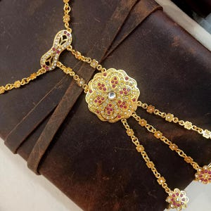 May include: A gold-toned hand chain with a floral design. The chain features a central, ornate flower pendant with red and clear gemstones. Delicate chains extend to finger rings, and the piece is set against a brown leather surface.