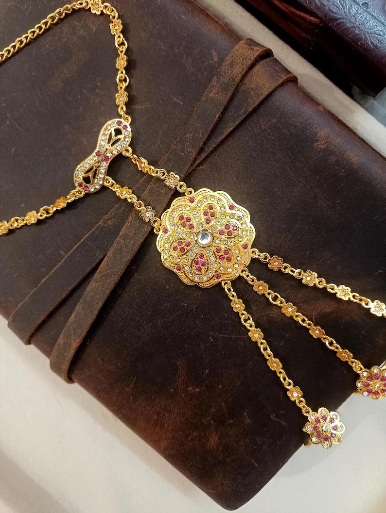May include: A gold-toned hand chain with a floral design. The chain features a central, ornate flower pendant with red and clear gemstones. Delicate chains extend to finger rings, and the piece is set against a brown leather surface.