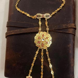 May include: A gold-colored hand chain bracelet with a floral design. The bracelet features a chain that goes around the wrist and connects to three finger rings. The bracelet is adorned with red and clear gemstones.
