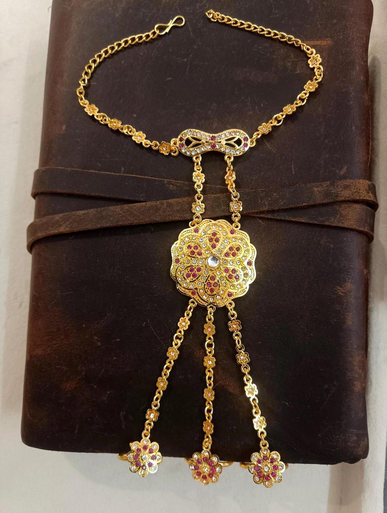 May include: A gold-colored hand chain bracelet with a floral design. The bracelet features a chain that goes around the wrist and connects to three finger rings. The bracelet is adorned with red and clear gemstones.