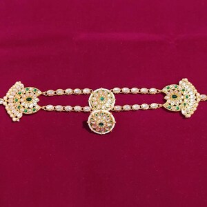 May include: Gold-coloured headpiece with detailed floral and geometric patterns. The piece features clear and green gemstones, with a central pendant and side accents. The headpiece is displayed against a deep red background.