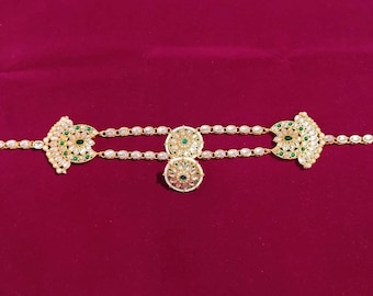 May include: Gold-coloured headpiece with detailed floral and geometric patterns. The piece features clear and green gemstones, with a central pendant and side accents. The headpiece is displayed against a deep red background.