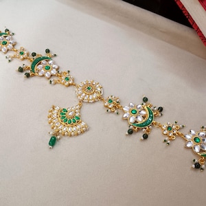 May include: Gold-toned headpiece with green and white accents. The design features floral and crescent moon shapes, adorned with green and white gemstones. A central pendant and dangling green beads complete the ornate look.