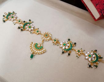 May include: Gold-coloured headpiece with green and white detailing. The design incorporates floral and crescent moon motifs, embellished with green and white gemstones. A central pendant and dangling green beads complete the ornate look.