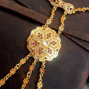 May include: Gold-toned hand chain jewelry with a floral design. The central piece features a large, ornate flower shape with red and clear gemstones. Delicate chains extend to the finger and wrist, creating an elegant accessory.