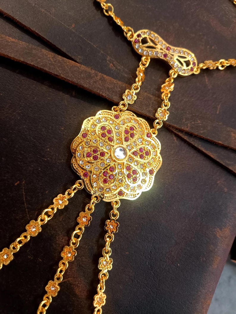 May include: Gold-toned hand chain jewelry with a floral design. The central piece features a large, ornate flower shape with red and clear gemstones. Delicate chains extend to the finger and wrist, creating an elegant accessory.
