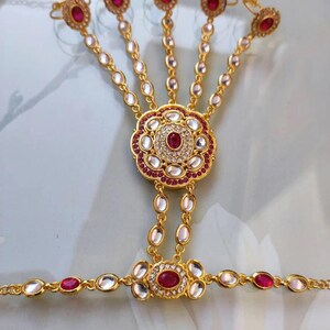 Pu&ograve; includere: Gioiello per mano dorato con un motivo floreale centrale con una grande gemma rossa circondata da pietre bianche e rosse pi&ugrave; piccole. Il pezzo si collega a cinque anelli per le dita tramite delicate catene, ognuno con una gemma rossa.